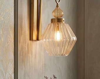 May include: A gold-toned wall sconce with a clear, faceted glass shade. The light fixture has a curved arm and a decorative finial. The sconce is mounted on a light-colored wall, with a small vase and clock on a nearby table.