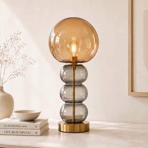 May include: A table lamp featuring a large amber-tinted spherical glass shade atop a vertical stack of three smaller, smoke-grey glass spheres. The lamp has a central brass-coloured metal rod and a circular brass-coloured base with an internal bulb.