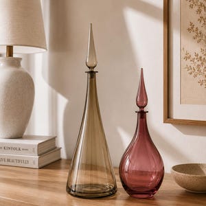 May include: Two glass decanters with pointed stoppers. The taller decanter is a translucent amber colour with a conical base, while the shorter decanter is a translucent plum colour with a rounded, bulbous base. Both feature smooth glass.