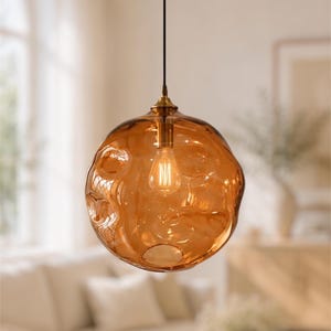 Könnte beinhalten: Eine kugelförmige Pendelleuchte mit einem transparenten, bernsteinfarbenen Glasschirm, der eine unregelmäßige, gewellte Oberflächenstruktur aufweist. Die Glaskugel hängt an einem schwarzen Kabel an einer messingfarbenen Halterung mit Leuchtmittel.