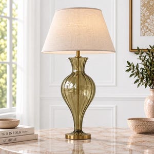 Puede incluir: Una lámpara de mesa con una base de vidrio acanalado en color verde oliva translúcido, cuello de metal en tono latón y base circular. La lámpara tiene una pantalla cónica de tela color blanco roto que emite una luz suave y difusa.