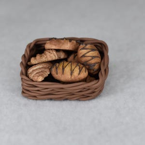 May include: A small brown woven basket containing various miniature baked goods. The basket holds croissants and bread rolls, some with chocolate drizzle. The pastries are light brown with darker brown details. The basket is square and has a woven texture.