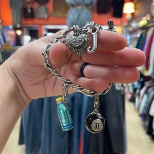 Op de afbeelding: Een zilverkleurige schakelarmband met een hartvormige bedel bezet met doorzichtige strass-steentjes en de tekst Juicy Couture, een letter J-bedel, een turquoise flesbedel met een gele dop en een zwarte acht-ball-bedel.