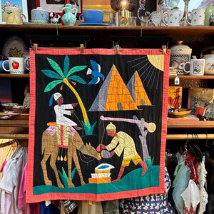 May include: A rectangular fabric wall hanging with a black background and a coral-colored border. The scene features a person riding a brown camel under a palm tree, a person standing near a well, two yellow pyramids, and a blue bird in the sky.