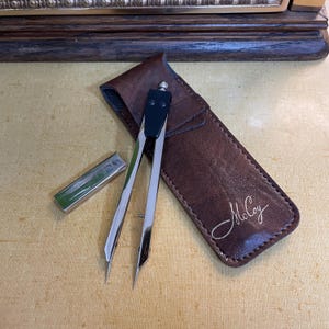 Puede incluir: Un compás de dibujo plateado y negro con un estuche de cuero marrón. Una pequeña regla plateada está al lado del compás. El estuche de cuero lleva el nombre "McCoy".