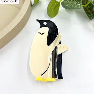 Puede incluir: Una pinza para el pelo de pingüino en blanco y negro con patas amarillas. El pingüino está mirando hacia la derecha y tiene un pico y ojos negros.