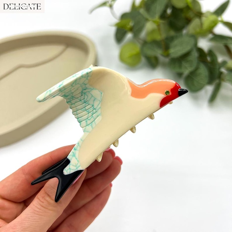 May include: A decorative bird-shaped hair clip. The clip features a cream-colored body with coral and red accents on the head and a turquoise wing design. The tail is black. The word "DELICATE" is in the upper left corner.