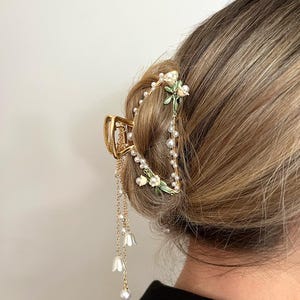 May include: Gold-toned hair claw clip with floral and pearl accents. The clip features a decorative design with small white flowers, faux pearls, and dangling pearl strands. The word "DELICATE" is printed at the top left.