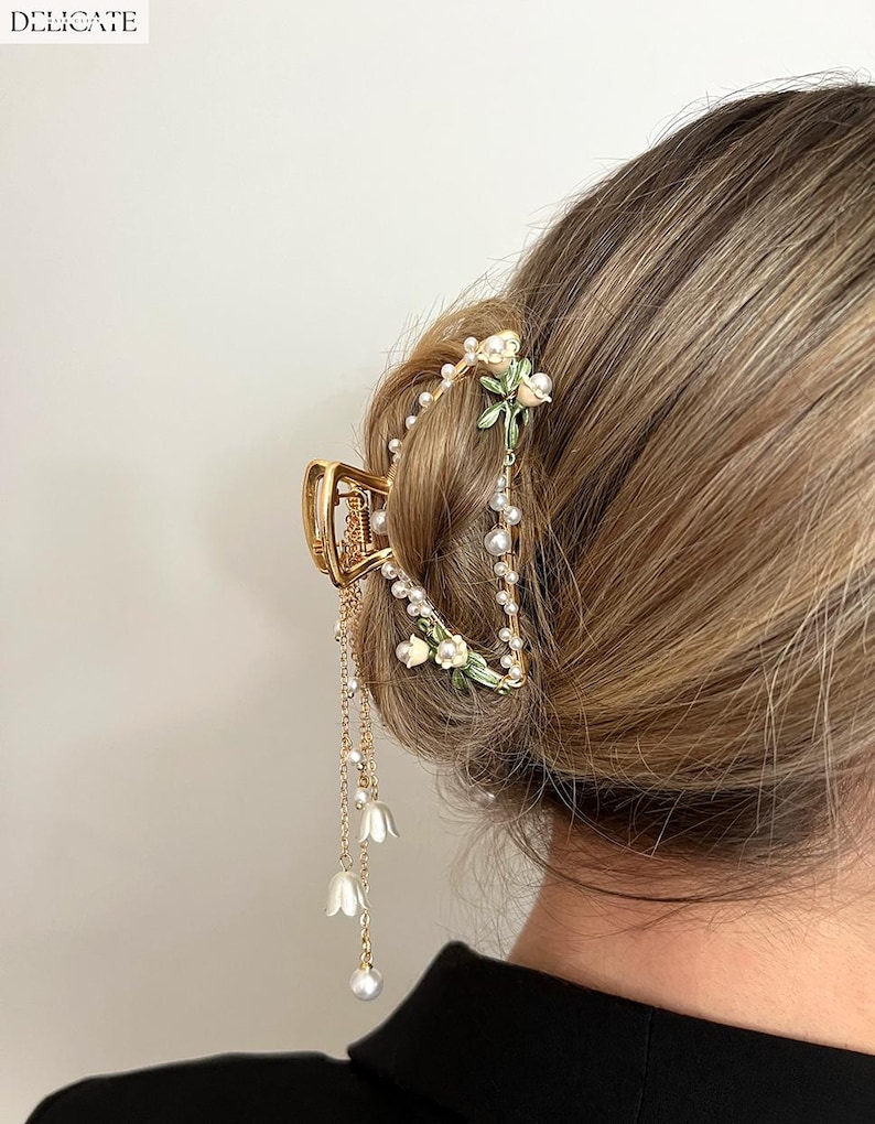 May include: Gold-toned hair claw clip with floral and pearl accents. The clip features a decorative design with small white flowers, faux pearls, and dangling pearl strands. The word "DELICATE" is printed at the top left.