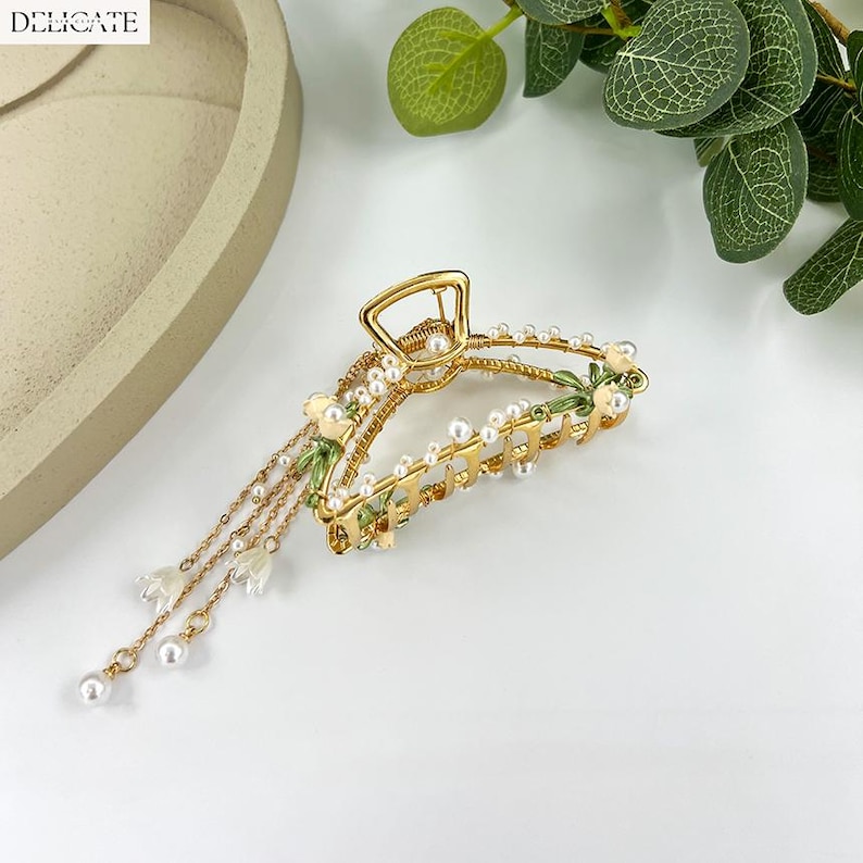 May include: Gold-toned hair claw clip with a floral design. The clip features small white pearls, green enamel accents, and dangling chains with pearl and flower charms. The word "DELICATE" is printed in white in the upper left corner.