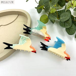May include: Three bird-shaped hair clips in a variety of colors, including blue, orange, and cream. Each clip has a black tail and beak, with the word "DELICATE" in the upper left corner. The clips are on a white surface.