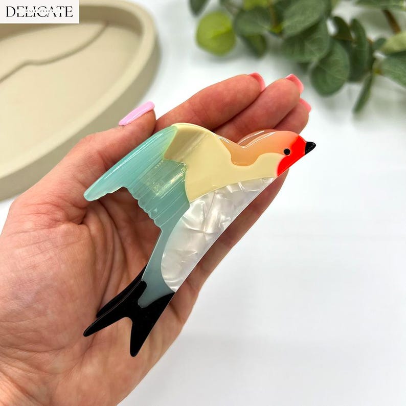 May include: A colorful bird-shaped brooch with a red head, orange beak, and blue wings. The brooch has a pearlescent white body and black tail feathers. The word "DELICATE" is printed in white in the upper left corner.