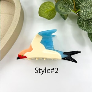 May include: Three colorful hair claw clips in the shape of birds. Style #1 features a bird with orange, yellow, blue, and white accents. Style #2 has blue, orange, and cream colors. Style #3 has green, orange, and cream colors. Each clip has a black tail.
