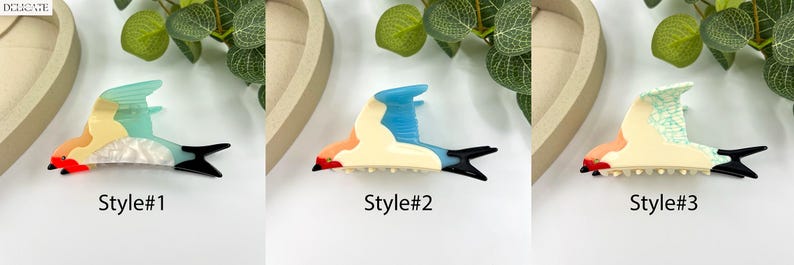 May include: Three colorful hair claw clips in the shape of birds. Style #1 features a bird with orange, yellow, blue, and white accents. Style #2 has blue, orange, and cream colors. Style #3 has green, orange, and cream colors. Each clip has a black tail.
