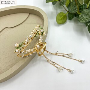 May include: A gold-toned hair claw clip with a floral design. The clip features white faux pearls, green enamel accents, and dangling chains with small white flower charms and pearl beads. The word "DELICATE" is printed at the top.