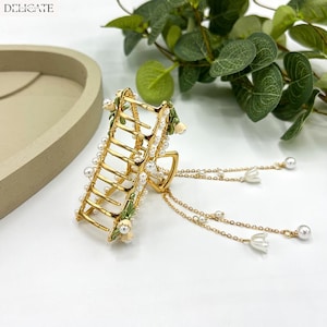 May include: Gold-toned hair claw clip with pearl accents and floral details. The clip features a ladder-like design adorned with small pearls and delicate white flowers. Dangling chains with pearls and flower charms add a touch of elegance. The word "DELICATE" is printed at the top.