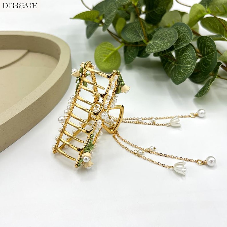 May include: Gold-toned hair claw clip with pearl accents and floral details. The clip features a ladder-like design adorned with small pearls and delicate white flowers. Dangling chains with pearls and flower charms add a touch of elegance. The word "DELICATE" is printed at the top.