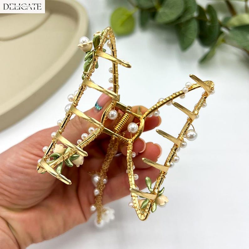 May include: Gold-toned metal hair claw clip with faux pearls and floral accents. The clip features a delicate design with small white flowers and green leaves. The word "DELICATE" is printed in the upper left corner.