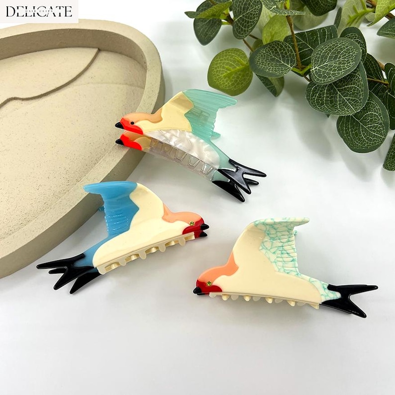 May include: Three colorful bird-shaped hair clips. Each clip features a unique color scheme, including blue, peach, and cream, with black tail feathers and red accents. The word "DELICATE" is in the top left corner.