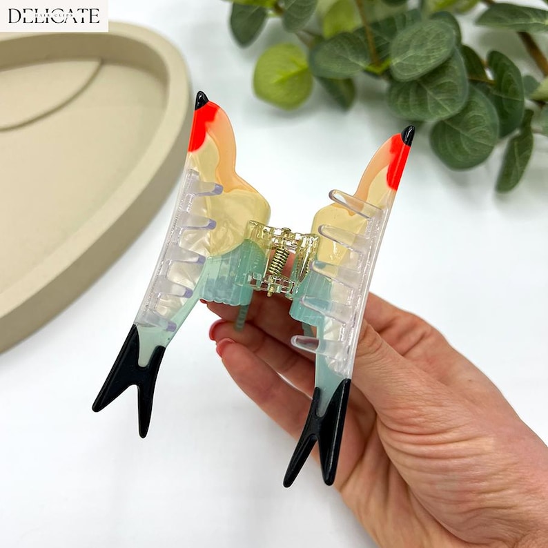 May include: A colorful hair claw clip with a butterfly design. The clip features a clear body with red, yellow, and blue accents. The tips are black, and the word "DELICATE" is in the top left corner.