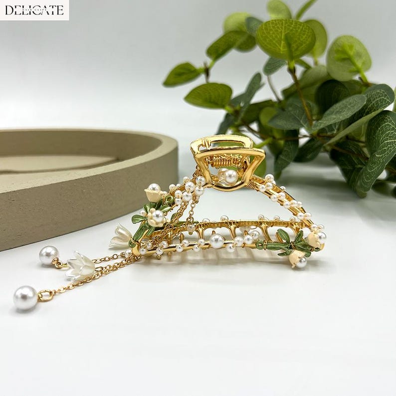 May include: Gold-toned hair claw clip adorned with faux pearls, white flowers, and green leaves. The clip features a delicate design with dangling pearl accents. The word "DELICATE" is printed in white in the upper left corner.