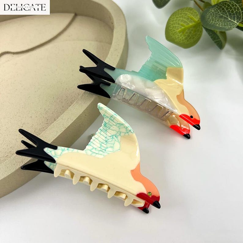 May include: Two bird-shaped hair clips. The clips feature a colorful design with black, red, cream, and teal accents. The word "DELICATE" is in the top left corner. These decorative hair accessories are designed to hold hair in place.