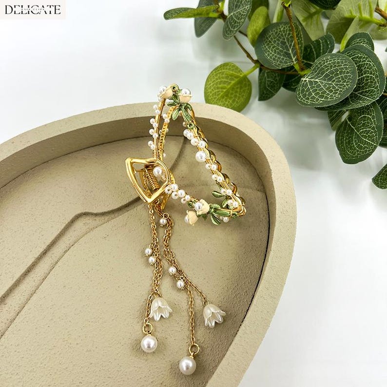 May include: Gold-toned hair claw clip with a floral design. The clip features a heart-shaped clasp and dangling chains adorned with pearls and small white flower charms. The clip is decorated with pearls and green and white floral accents.