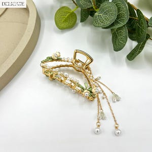 May include: Gold-toned hair claw clip with pearl and floral accents. The clip features a delicate design with small white flowers and dangling pearl charms. The word "DELICATE" is printed in white in the upper left corner.