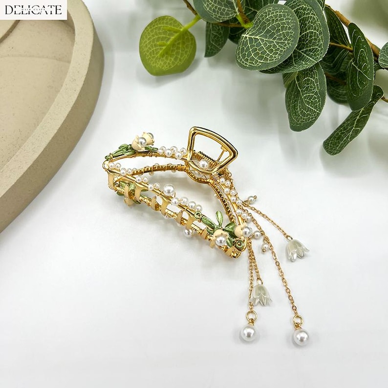 May include: Gold-toned hair claw clip with pearl and floral accents. The clip features a delicate design with small white flowers and dangling pearl charms. The word "DELICATE" is printed in white in the upper left corner.