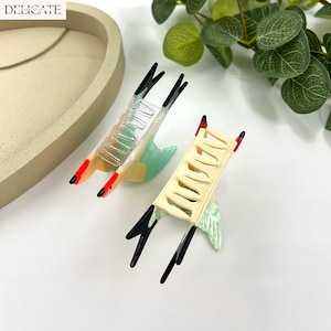 May include: Two decorative hair clips with a bird-like design. The clips feature a cream-colored base, black accents, and red tips. One clip is clear, and the other is cream-colored. The clips are set against a white background with greenery.