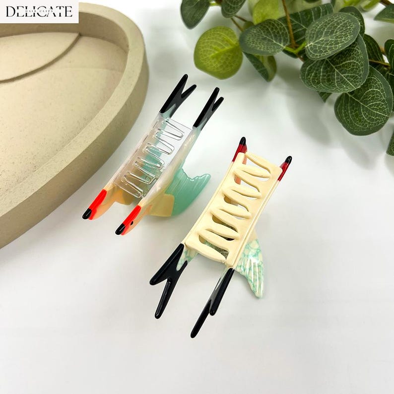May include: Two decorative hair clips with a bird-like design. The clips feature a cream-colored base, black accents, and red tips. One clip is clear, and the other is cream-colored. The clips are set against a white background with greenery.