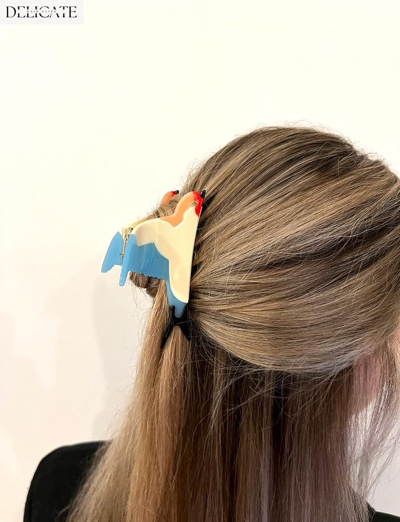 May include: A colorful hair clip in the shape of a bird, featuring shades of blue, cream, orange, and red. The clip is attached to light brown hair. The word "DELICATE" is printed in white at the top left.