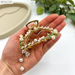 May include: Gold-toned hair claw clip adorned with faux pearls and white floral accents. The clip features a delicate design with green painted leaves and dangling pearl charms, adding a touch of elegance. The word "DELICATE" is printed on the top left.