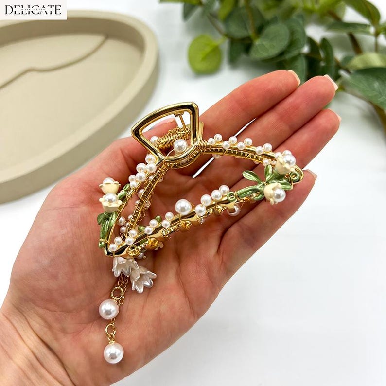 May include: Gold-toned hair claw clip adorned with faux pearls and white floral accents. The clip features a delicate design with green painted leaves and dangling pearl charms, adding a touch of elegance. The word "DELICATE" is printed on the top left.