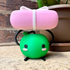May include: A whimsical, 3D-printed sculpture featuring a green, round character with black eyes and pink cheeks, supported by a black stand. A pink, rectangular object with a white bow and band sits atop the stand. The sculpture is set against a brick wall and a potted plant.