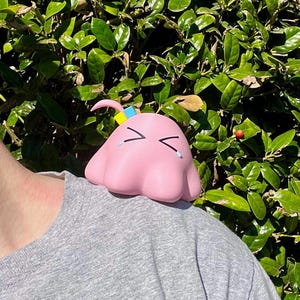 May include: A pink, blob-shaped character with closed eyes and teardrops, perched on a gray shirt. The character has a small, curved pink antenna with yellow and blue accents. The background features green foliage.