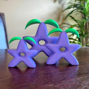 May include: Three purple star-shaped decorative objects with green leaf accents. Each star has a central hole. The objects are arranged on a dark wooden surface, with a blurred background.