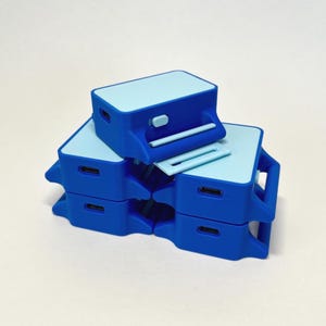 May include: A stack of four rectangular blue plastic devices with light blue top panels. Each device features a small rectangular port on the side, a raised horizontal bar, and a small protruding button on the front face of the main body.