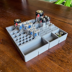 May include: A gray battery organizer with various compartments and slots, holding different sizes of batteries, including AA, AAA, C, D, and 9-volt batteries. The organizer also has a section for button cell batteries and coins. The batteries are of the Energizer brand.