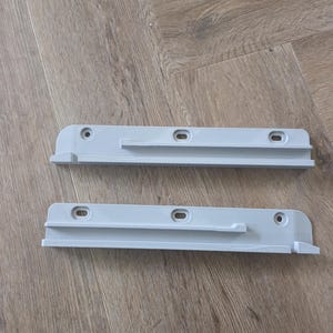 Op de afbeelding: Twee lichtgrijze plastic plankdragers. Elke drager heeft een rechthoekige vorm met een lip en drie voorgeboorde gaten voor montage. De dragers zijn ontworpen om planken te ondersteunen.