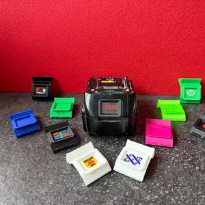 Könnte beinhalten: Ein schwarzes elektronisches Gerät mit dem Text "HWM" und mehreren bunten Kunststoffkomponenten. Die Komponenten sind in verschiedenen Farben, darunter Blau, Grün, Pink und Weiß, und weisen unterschiedliche Designs auf.
