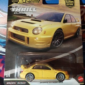 Op de afbeelding: Een gele Hot Wheels-gietijzeren auto op een zwart-gele kaart. De kaart bevat de tekst "Hot Wheels Premium Thrill Climbers" en "10 Car Culture". De auto is een Subaru Impreza WRX.