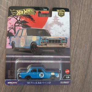 Peut inclure: Une voiture miniature Hot Wheels bleue, modèle '64 Prince Skyline GT, avec le numéro 39 sur le côté. L'emballage présente une illustration d'inspiration japonaise avec un soleil rouge et une pagode. La voiture fait partie de la série Car Culture.
