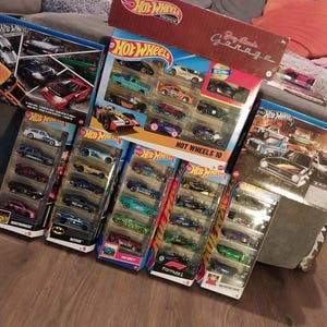 May include: A collection of Hot Wheels toy car sets, featuring various car models in different colors. The sets are packaged in colorful boxes and blister packs, with some sets labeled "Hot Wheels Premium" and "Day Lane's Garage".