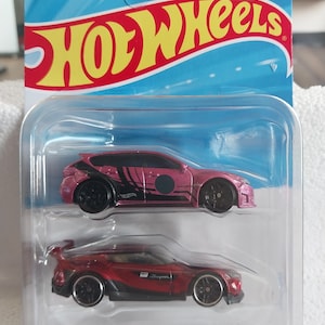 Puede incluir: Un paquete doble de Hot Wheels con un coche rosa con detalles negros y un coche deportivo rojo. El embalaje incluye el logotipo de Hot Wheels en amarillo y rojo. Los coches están en una caja de plástico transparente.
