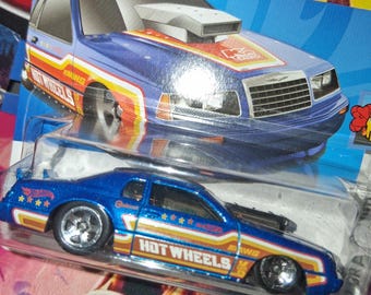 May include: A blue Hot Wheels toy car with a white, orange, and yellow stripe design. The car has the Hot Wheels logo and text on the side. The car is in a clear plastic package with a blue and yellow background.