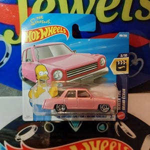 May include: A pink Hot Wheels die-cast car of The Simpsons Family Car, displayed on its original packaging. The packaging is blue and features Homer Simpson. The car is number 108 of 250 in the series.