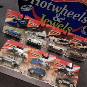 Może przedstawiać: Kolekcja siedmiu odlewanych modeli samochodów Matchbox w oryginalnych opakowaniach typu blister, w tym niebieski kabriolet, białe coupe, biały kabriolet, beżowe coupe, biały pickup, niebieski SUV i zielona terenówka, ustawione przed szyldem.