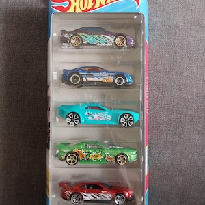 Op de afbeelding: Een vijfpack Hot Wheels speelgoedauto's in een doorzichtige plastic verpakking. De auto's zijn in verschillende kleuren, waaronder paars, blauw, turquoise, groen en rood. Elke auto heeft unieke graphics en ontwerpen. De verpakking heeft het Hot Wheels-logo en de tekst "HW DRIFT".