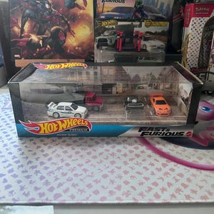 May include: A Hot Wheels Premium set featuring four miniature Fast & Furious cars. The set includes a white car, a red truck, a black car, and an orange car, all displayed in a clear plastic case with the Hot Wheels logo.